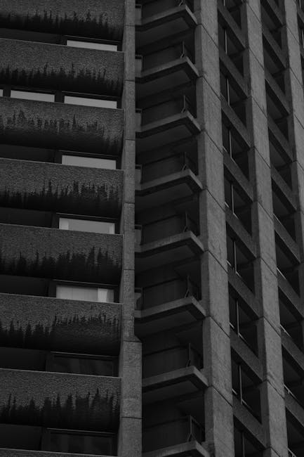 The image depicts a close-up view of a modern multi-storey residential building with a concrete facade, featuring multiple balconies on the left side and a series of vertically aligned windows and small balconies on the right side. The balconies on the left have dark railings and some greenery visible on the ledges, possibly potted plants or shrubs, while the right side shows a repeating pattern of small balconies with glass doors, creating a geometric architectural pattern. The building's dark exterior contrasts with the lighter sky visible in the background, suggesting overcast weather. This architectural detail highlights the urban environment typical of a city like Barbican, which may be relevant to house removals or home relocation services carried out by companies such as Man with Van Barbican, especially during packing, furniture transport, or loading processes involved in moving into or out of high-rise apartments.