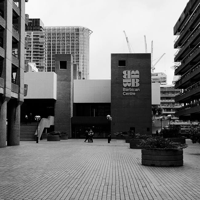 A black and white photograph showing the exterior of the Barbican Centre on Aldersgate Street, with a paved pedestrian area in the foreground. The building features a modern architectural style with two prominent brick towers displaying the 'Barbican Centre' logo and large windows. Surrounding the entrance are several planters with small trees and bushes, and a few pedestrians are seen walking or standing nearby. To the left, part of a multi-storey building and some scaffolding cranes are visible in the background, indicating ongoing construction or development in the area. The scene appears to be during daylight hours, with natural light illuminating the urban environment, providing context for house removals and furniture transport activities related to home relocation projects, supported by companies like Man with Van Barbican.