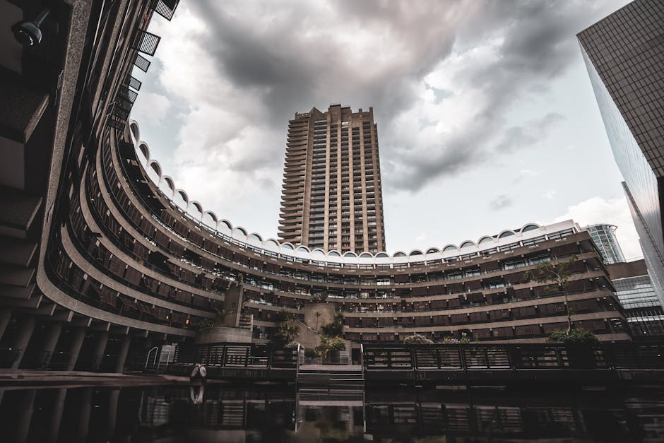 The image depicts a close-up view of a modern multi-storey residential building with a concrete facade, featuring multiple balconies on the left side and a series of vertically aligned windows and small balconies on the right side. The balconies on the left have dark railings and some greenery visible on the ledges, possibly potted plants or shrubs, while the right side shows a repeating pattern of small balconies with glass doors, creating a geometric architectural pattern. The building's dark exterior contrasts with the lighter sky visible in the background, suggesting overcast weather. This architectural detail highlights the urban environment typical of a city like Barbican, which may be relevant to house removals or home relocation services carried out by companies such as Man with Van Barbican, especially during packing, furniture transport, or loading processes involved in moving into or out of high-rise apartments.