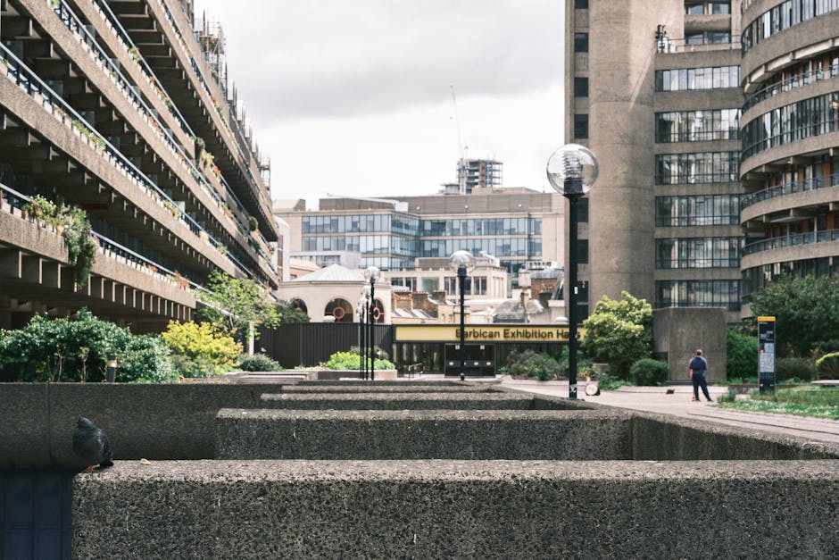 A daytime exterior view of the Barbican Exhibition Hall entrance located within a central courtyard surrounded by tall, multi-storey residential and commercial buildings. The courtyard features stone planters with green shrubs and small trees, and several modern, spherical streetlamps mounted on black poles. In the foreground, there are large, rectangular stone or concrete blocks forming part of the courtyard landscape. Toward the center, a person dressed in dark clothing is walking near a black information board, while another individual is visible in the distance. The scene is well-lit with natural light, and the overall environment suggests an urban setting suitable for house removals and furniture transport logistics in central London. The image emphasizes the building's architectural features and the open space used during moving or loading processes, aligning with services offered by Man with Van Barbican for house removals in the EC2Y area.