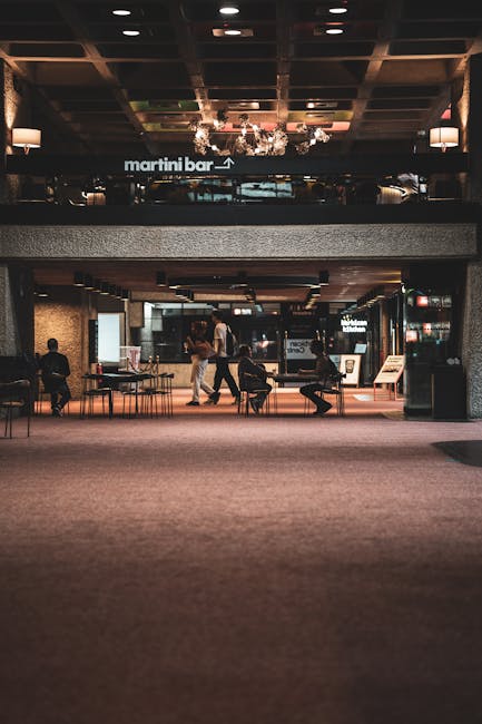 The image shows the interior of a public venue with a spacious, carpeted floor area and a high ceiling featuring a grid-style wooden design with built-in lighting. In the background, there is a sign indicating the direction to the Martini Bar. Several people are seen inside, some walking and others seated at tables, with some engaged in conversation or waiting. The environment is illuminated with warm, ambient lighting, and the walls have textured finishes. To the right, a small kiosk or sign stand is visible. The scene depicts a typical setting for casual socialising or waiting, relevant to the context of venue relocations and moving services coordinated by Man with Van Barbican, especially when planning transport or reallocation within large public spaces like the Barbican Centre.