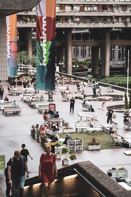 A daytime exterior view of the Barbican Exhibition Hall entrance located within a central courtyard surrounded by tall, multi-storey residential and commercial buildings. The courtyard features stone planters with green shrubs and small trees, and several modern, spherical streetlamps mounted on black poles. In the foreground, there are large, rectangular stone or concrete blocks forming part of the courtyard landscape. Toward the center, a person dressed in dark clothing is walking near a black information board, while another individual is visible in the distance. The scene is well-lit with natural light, and the overall environment suggests an urban setting suitable for house removals and furniture transport logistics in central London. The image emphasizes the building's architectural features and the open space used during moving or loading processes, aligning with services offered by Man with Van Barbican for house removals in the EC2Y area.