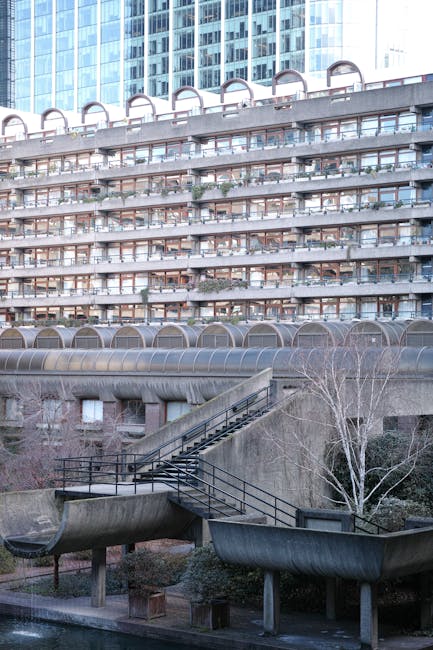 A multi-storey residential building with numerous balconies featuring glass railings and curved metal coverings, standing behind an outdoor concrete staircase with black metal railings, leading from a paved area at the base. In the foreground, two large, industrial-style plastic ducts are supported by concrete bases, with leafless trees and shrubs nearby. The scene is captured during daylight with overcast weather, and the building's exterior shows a modern, functional design typical of urban housing. The image, associated with local house removal and furniture disposal services near Barbican, highlights the urban environment suitable for furniture transport and home relocation logistics handled by Man with Van Barbican.