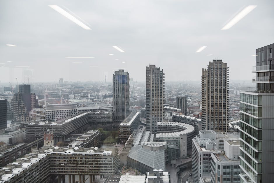 A high-angle view from inside a modern building showing a cityscape with multiple high-rise office buildings and residential towers, some with glass facades. The foreground features a building with a rooftop terrace and white exterior, adjacent to a parking area. In the background, the skyline extends into a hazy, overcast sky, with construction cranes visible among the urban landscape. The scene is lit by diffuse natural light, highlighting the structural details of the buildings. This image reflects the urban environment in which house removals or furniture transport services by Man with Van Barbican might operate, supporting the context of city-based relocation and logistical planning within the City of London.