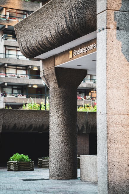 A close-up view of the exterior of a commercial building featuring a large, textured cylindrical column made of concrete supporting an overhanging structure with a smooth, rounded edge. The building's