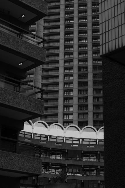 Photograph taken from a lower angle showing the facades of modern residential buildings with multiple floors, balconies, and large windows. The buildings are constructed with dark brick and concrete, and the image captures a section of a courtyard or communal area with a curved, white-roofed structure visible at the center. In the foreground, parts of exterior walls and balconies frame the view, which is illuminated by natural daylight. The scene reflects an urban setting suitable for house removals and home relocation services, illustrating the environment where a professional movers company like Man with Van Barbican might operate when assisting clients with furniture transport and packing within apartment complexes such as the Golden Lane Estate.