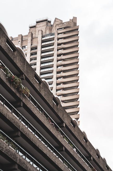 The image displays a close-up view of a high-rise residential building's exterior, highlighting the concrete balcony structures and window arrangements. The building features multiple floors with protruding balconies, some of which are equipped with metal railings and small potted plants. The facade appears weathered with visible streaks and streaking marks on the concrete surfaces, suggesting exposure to the elements. The photograph captures the building in natural light, emphasizing the stark, utilitarian design typical of urban housing complexes. The perspective focuses on the vertical alignment of the balconies and windows, providing a sense of the building's height and architectural style. This type of image could relate to the context of house or flat removals, illustrating a typical urban setting where furniture and belongings are transported during home relocation or moving services. Man with Van Barbican's expertise in furniture transport and packing would be relevant to such a setting, especially when navigating high-rise buildings like this one.