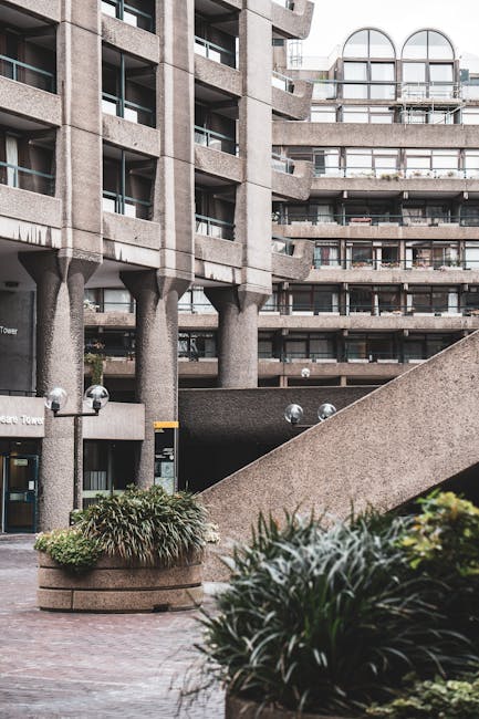 Close-up view of the exterior of a modern building with a textured concrete column and canopy, featuring a wooden sign with the partial name 'Shakespeare' attached to the building facade. In the backg