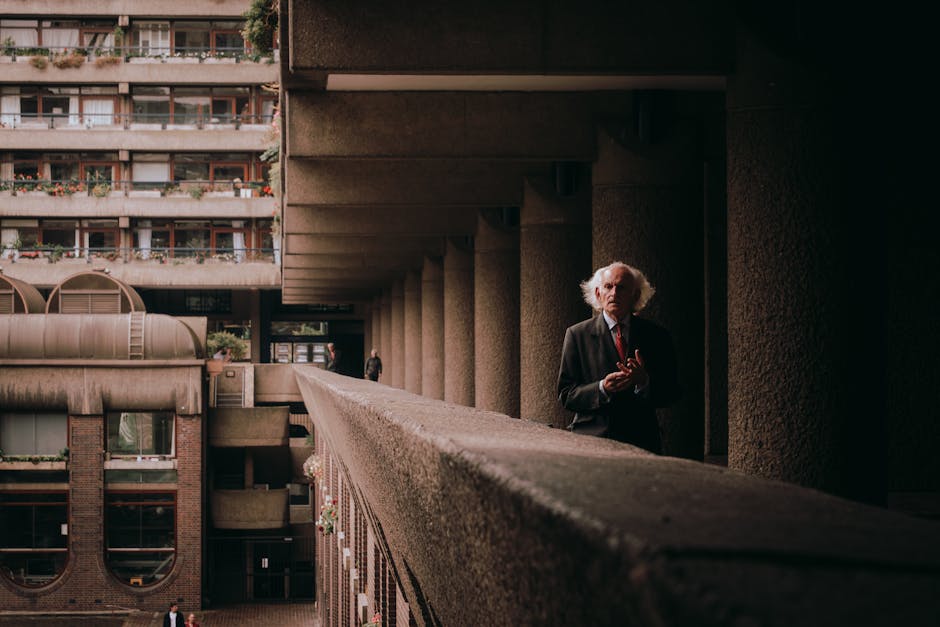 A man with white, disheveled hair and glasses standing on a concrete balcony outside a multi-storey residential building, which features balconies with potted plants and glass doors. The man is wearing a dark suit jacket over a white shirt with a red tie, and appears to be holding or adjusting something in his hands. The balcony is supported by large concrete pillars, with a dark shaded area underneath. Behind the building, a person in dark clothing is seen walking in the background. The scene is lit with natural daylight, highlighting the textures of the concrete and brickwork. This image depicts the process of packing and moving, set within an urban residential environment, and is associated with house removals and furniture transport services provided by Man with Van Barbican.