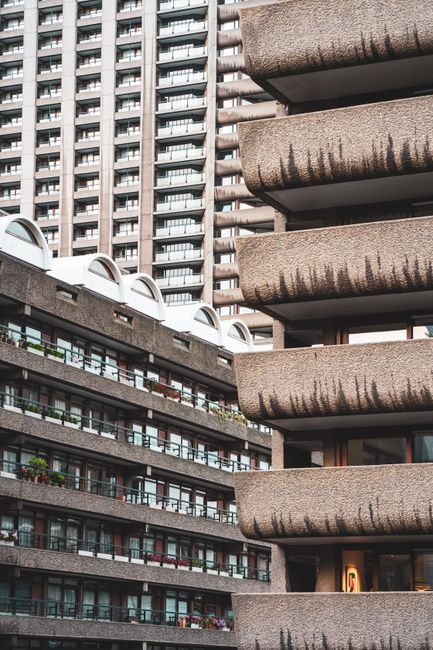 Image depicting the exterior of two intersecting residential buildings with multiple balconies, characteristic of the Barbican estate. The building on the left features a concrete facade with a pattern of horizontal lines, and several balconies with glass railings and potted plants, while the building on the right showcases large, curved concrete balconies casting shadows on each other. The scene is taken during daylight with natural light illuminating the textured surfaces. This visual context relates to house removals and moving services, emphasizing the type of properties involved in the relocation process, as provided by Man with Van Barbican, situated within the context of furniture transport and loading activities associated with home relocation in listed Barbican buildings.