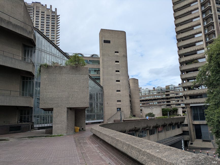 A wide outdoor view of a modern, brutalist-style building complex featuring concrete structures, including a tall, narrow tower with small rectangular windows, alongside lower sections with large glass panels. The scene shows an open paved area adjacent to the building, with a ramp leading down to a lower level and a portion of pavement being used for loading activities. There are no people or vehicles visible in movement, but the environment suggests a setting suitable for home relocation or furniture transport activities, as part of a professional removals service. The sky is partly cloudy, and there is greenery visible on the upper levels of the building. This image illustrates the type of building within which Man with Van Barbican might perform removals, emphasizing the concrete architecture and outdoor logistics involved in moving household items in an urban setting.