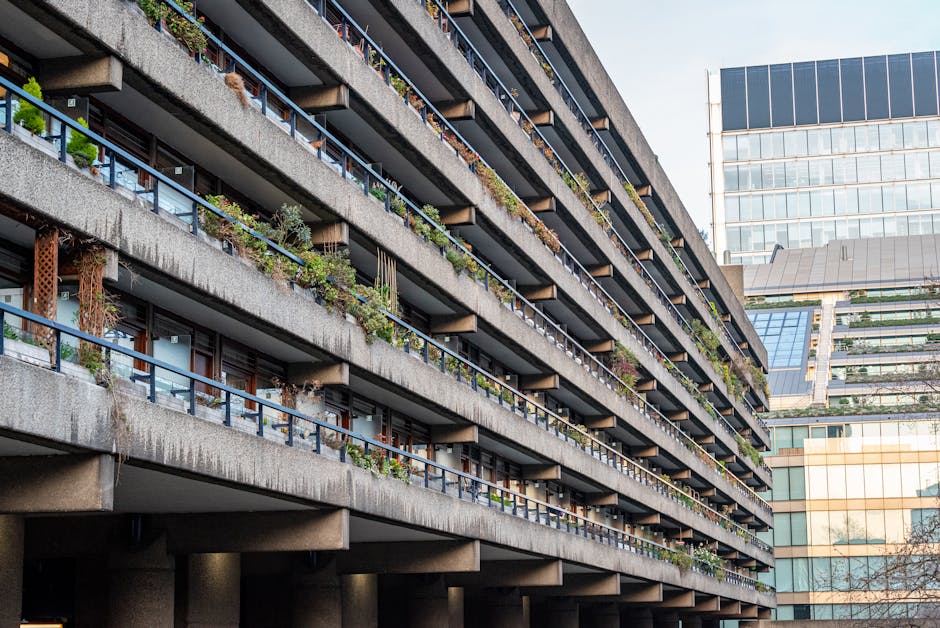 Image depicting the exterior of two intersecting residential buildings with multiple balconies, characteristic of the Barbican estate. The building on the left features a concrete facade with a pattern of horizontal lines, and several balconies with glass railings and potted plants, while the building on the right showcases large, curved concrete balconies casting shadows on each other. The scene is taken during daylight with natural light illuminating the textured surfaces. This visual context relates to house removals and moving services, emphasizing the type of properties involved in the relocation process, as provided by Man with Van Barbican, situated within the context of furniture transport and loading activities associated with home relocation in listed Barbican buildings.