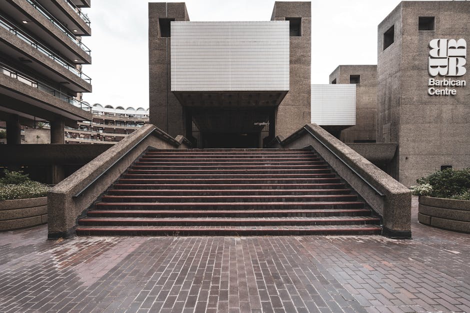 Exterior view of a concrete building identified as Barbican Centre with large white signage on the right side of the facade. In the foreground, a wide brick staircase with metal railings on both sides leads up to the entrance, which is partially shaded beneath an overhang. The stairs appear to be wet, possibly from recent rain, and are flanked by rounded concrete planters filled with greenery and small white flowers. To the left, part of a multi-storey residential or office building with balconies is visible, set against a cloudy sky. The scene captures a neutral, urban environment typical for house removals and furniture transport during a home relocation or moving process, with a focus on the external architecture and stairway access for loading and unloading furniture or boxes.
