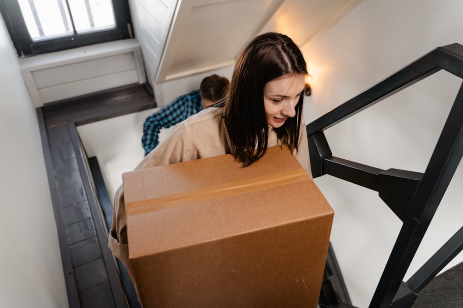 A woman with dark brown hair, wearing a beige coat, is carrying a large cardboard box up a narrow staircase in a residential setting. Behind her, a person in a blue and black checkered shirt is partially visible, assisting with the move. The staircase has black metal railings and dark wooden steps, with a white wall and a door at the top. Natural light from a nearby window illuminates the space, which features a small wall-mounted lamp. The scene depicts the physical process of home relocation, with careful handling of packed boxes and furniture, supported by a professional removals service such as Man with Van Barbican, as part of furniture transport and packing and moving operations.