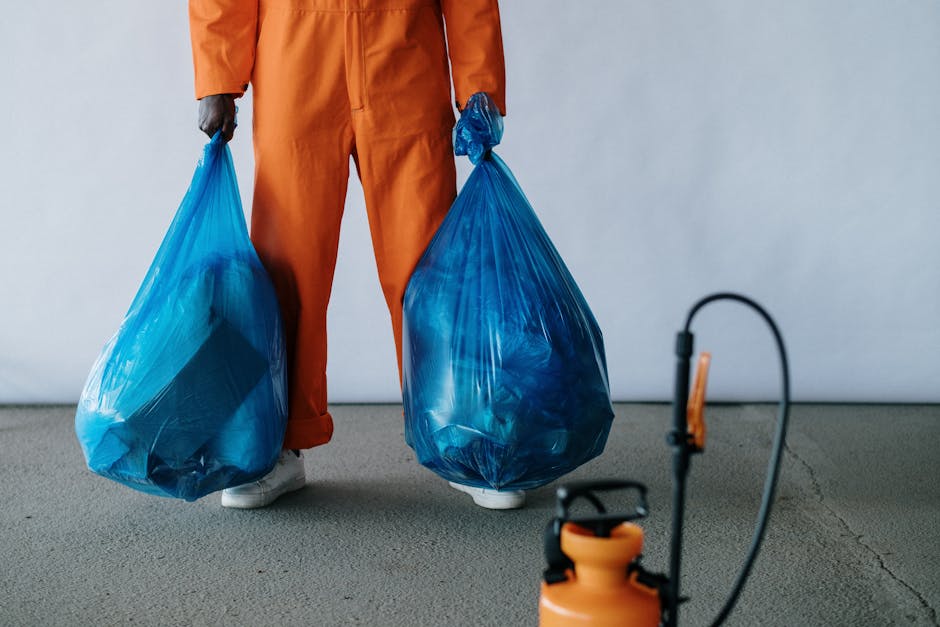 A person dressed in orange work overalls, wearing white shoes, is standing indoors on a grey carpeted floor, holding two large blue plastic rubbish bags filled with waste. One bag is held in each hand, with the person's lower half visible, focusing on their legs and hands. The background features a plain white wall, and nearby on the floor is an orange spray bottle with a black nozzle, connected to a small plastic tube. The scene depicts the process of packing and waste disposal during home relocation or furniture transport, aligned with house removal services provided by Man with Van Barbican. The image emphasizes the importance of proper waste management and packing materials, such as cardboard boxes and plastic bags, in compliance with removal waste rules in EC2, supporting efficient loading, transport, and disposal during moving logistics.